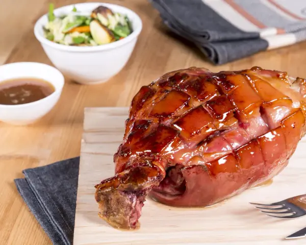 Glänzende, karamellisierte Schweinshaxe sous vide auf Holzbrett, daneben Schüssel mit Sauce und Salat.