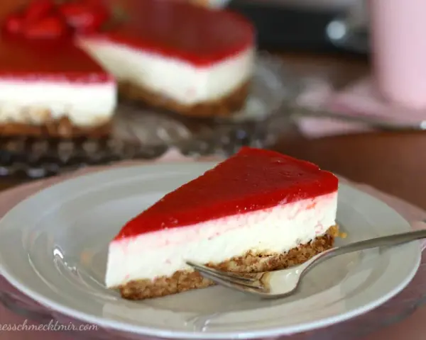 Ein Stück New York Cheesecake ohne Backen mit rotem Fruchttopping auf einem Teller, daneben ein ganzer Kuchen.