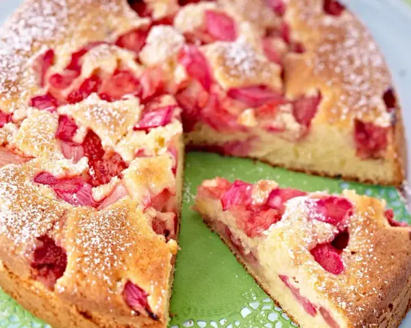 Ein saftiger Erdbeer Rhabarber Kuchen, bestäubt mit Puderzucker, wird auf einem Teller präsentiert. Ein Stück ist bereits herausgeschnitten.