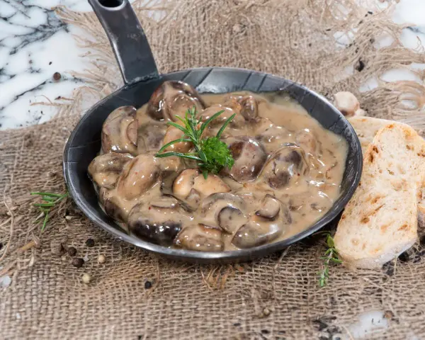 Herzhafte Champignon Rahmsauce in einer Pfanne, garniert mit Rosmarin, daneben knuspriges Brot und Pfefferkörner.