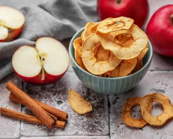 Leckere Apfelchips, die man ganz einfach selber machen kann, in einer Schüssel mit frischen Äpfeln und Zimtstangen.