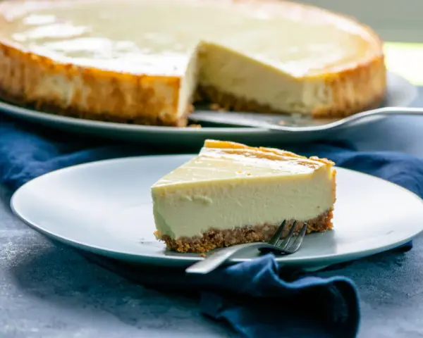 Ein Stück cremiger american cheesecake mit knusprigem Boden auf einem Teller, daneben ein ganzer Kuchen.