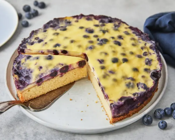 Ein saftiger Heidelbeerkuchen mit einer cremigen Vanillepudding-Schicht und frischen Heidelbeeren. Ein Stück wird gerade herausgeschnitten.