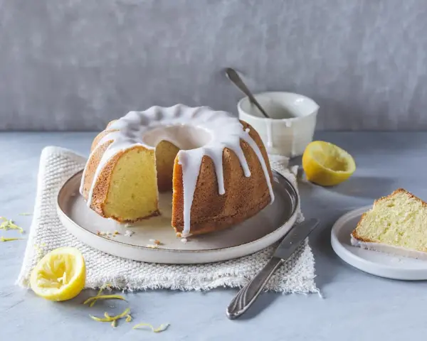 Ein saftiger Zitronenkuchen mit Zuckerguss, perfekt für ein Lemon Drizzle Cake Rezept. Daneben liegen frische Zitronen und ein Stück Kuchen.