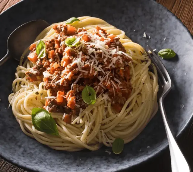 Ein Teller mit dampfenden Spaghetti Bolognese, garniert mit frischem Basilikum und Parmesan. Ein Löffel und eine Gabel liegen bereit.
