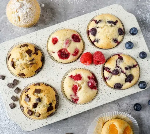 Leckeres Muffins Rezept: Schoko-, Himbeer- und Blaubeermuffins in einer Form, daneben frische Beeren und Schokostückchen.