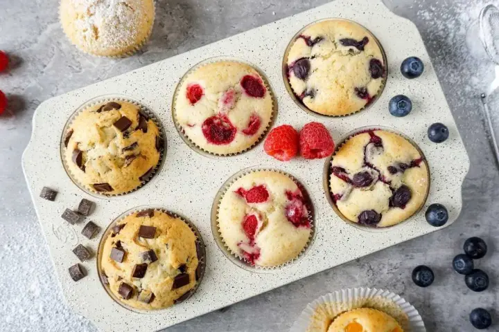 Leckeres Muffins Rezept: Schoko-, Himbeer- und Blaubeermuffins in einer Form, daneben frische Beeren und Schokostückchen.
