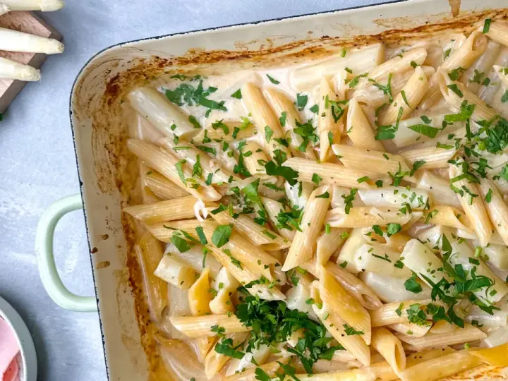 Cremige Spargel Pasta mit frischer Petersilie in einer Auflaufform. Dazu liegen Spargelstangen, Schinken und Parmesan bereit.