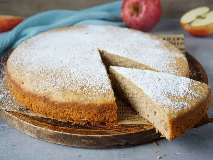Ein schneller, saftiger Kuchen mit Apfelmus, bestäubt mit Puderzucker. Ein Stück wird gerade herausgenommen.