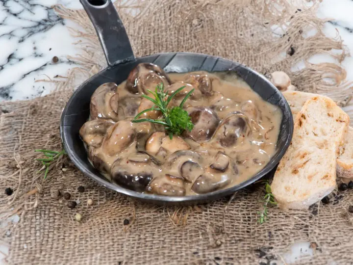 Herzhafte Champignon Rahmsauce in einer Pfanne, garniert mit Rosmarin, daneben knuspriges Brot und Pfefferkörner.