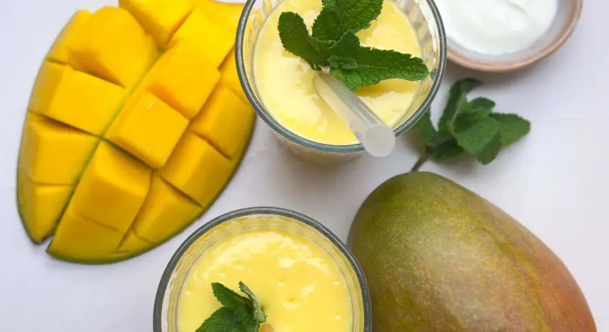 Zwei Gläser cremiger Mango Lassi, garniert mit Minze, neben frischen Mangos und einem Schälchen Joghurt.