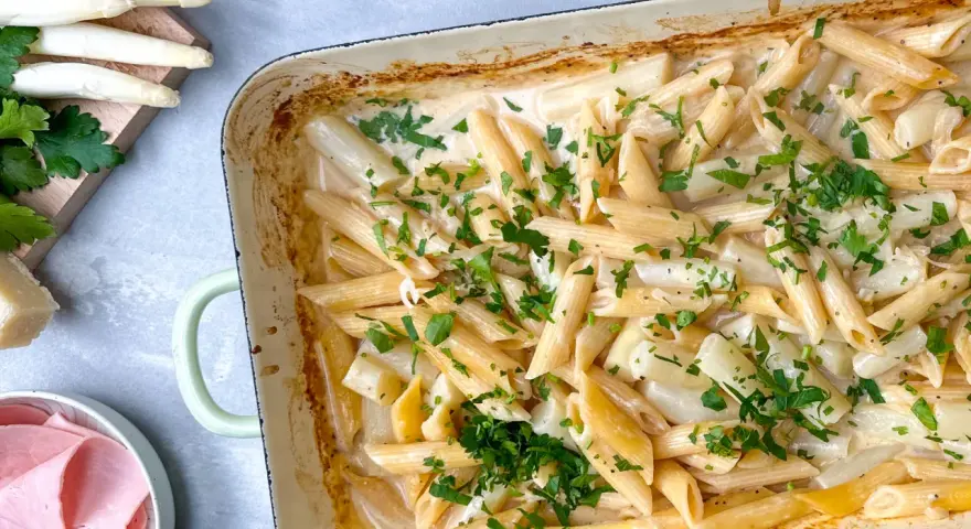 Cremige Spargel Pasta mit frischer Petersilie in einer Auflaufform. Dazu liegen Spargelstangen, Schinken und Parmesan bereit.