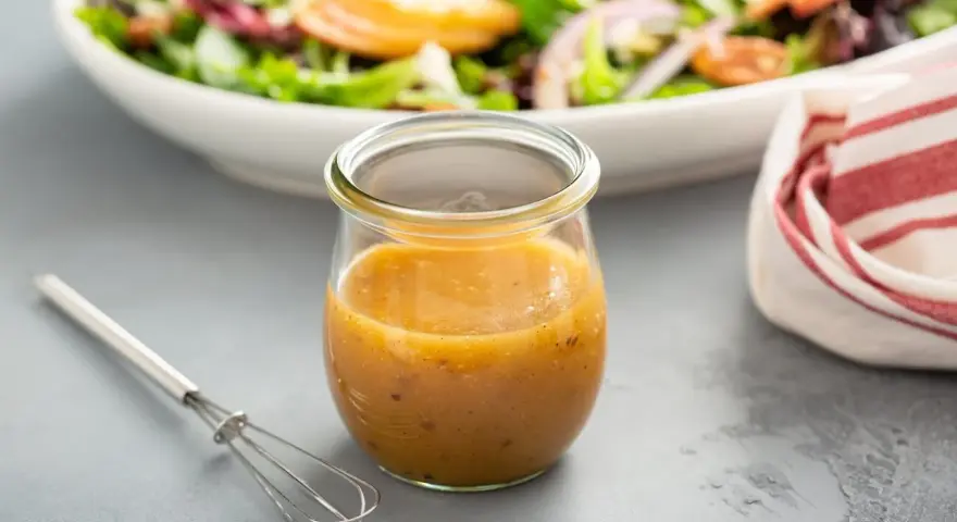 Ein Glas mit honig senf dressing steht neben einem kleinen Schneebesen. Im Hintergrund eine Schüssel mit Salat.