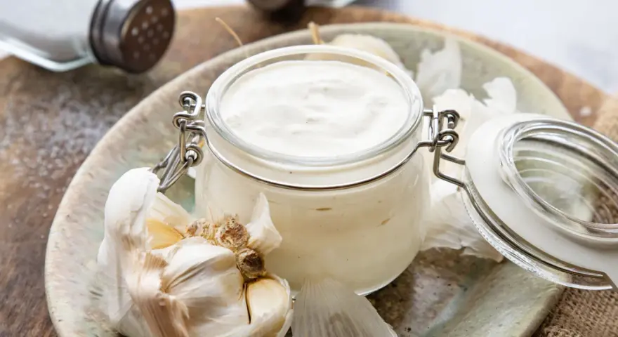 Frische Knoblauchsauce selber machen: cremige Sauce im Glas mit Knoblauchzehen und Salzstreuer im Hintergrund.