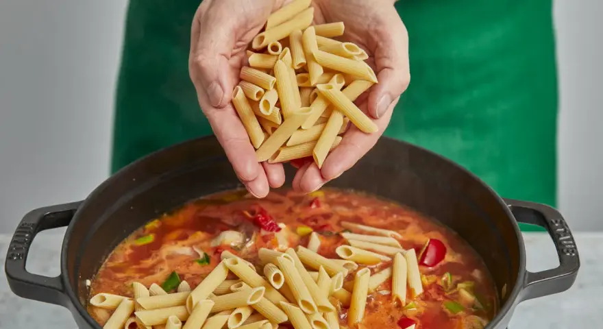 Hände geben Penne in einen Topf mit köchelnder Sauce für ein einfaches One-Pot-Chicken-Pasta-Gericht.