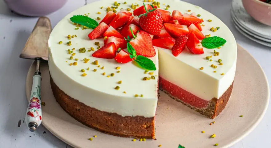 Erdbeer Joghurt Torte mit frischen Erdbeeren und Pistazien garniert. Ein Stück ist angeschnitten und zeigt die cremige Schicht.