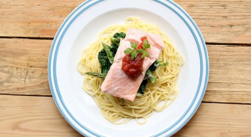 Ein Teller mit One Pot Lachs Pasta, garniert mit Petersilie und Tomatensauce, daneben Spargel und Spinat auf einem Holztisch.