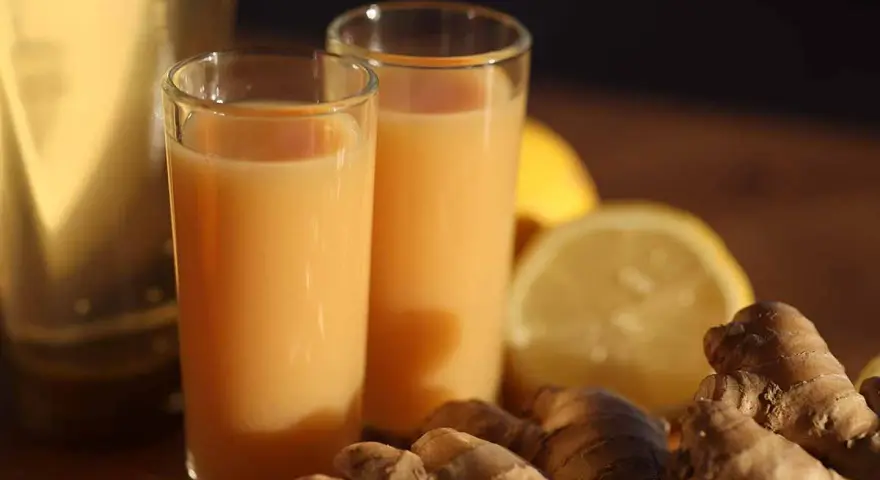 Zwei Gläser mit einem leuchtend orangen Ingwer Shot stehen neben frischem Ingwer und Zitronenscheiben.