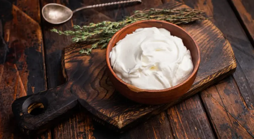 Cremiger, selbstgemachter Haferjoghurt in einer Holzschale, bereit zum Genießen. Ein Löffel und Kräuter liegen daneben.