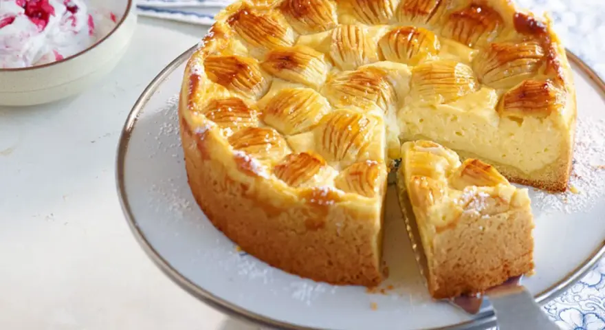 Ein köstlicher, schneller Apfelkuchen mit Schmand und Pudding, verziert mit Apfelscheiben und Puderzucker. Daneben eine Schüssel mit Beeren-Sahne.