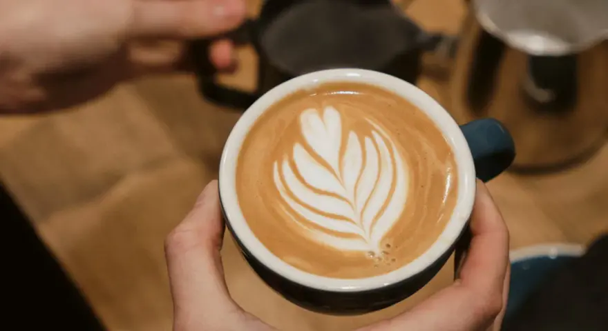 Cappuccino selber machen: Hände halten eine Tasse mit Latte Art. Im Hintergrund Milchkännchen und Sieb.
