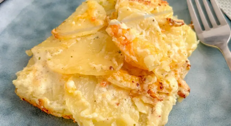 Ein Stück Kartoffelgratin einfach mit Sahne, goldbraun gebacken, auf einem blauen Teller. Daneben eine Gabel.