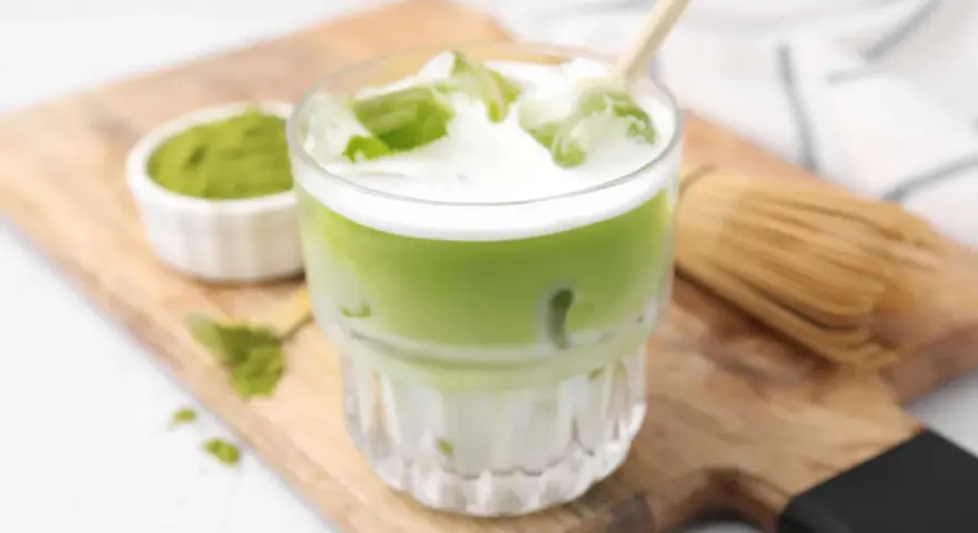 Erfrischender Matcha Latte mit Eis, Milch und einem Hauch von Starbucks-Rezept-Magie. Daneben eine Schale Matcha-Pulver und ein Chasen.