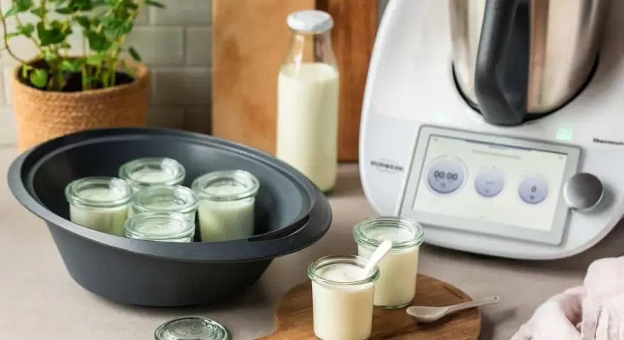 Kleine Gläser mit Joghurt in einem Dampfgarer, bereit, trinkjoghurt selber machen. Daneben ein Thermomix und Milchflasche.