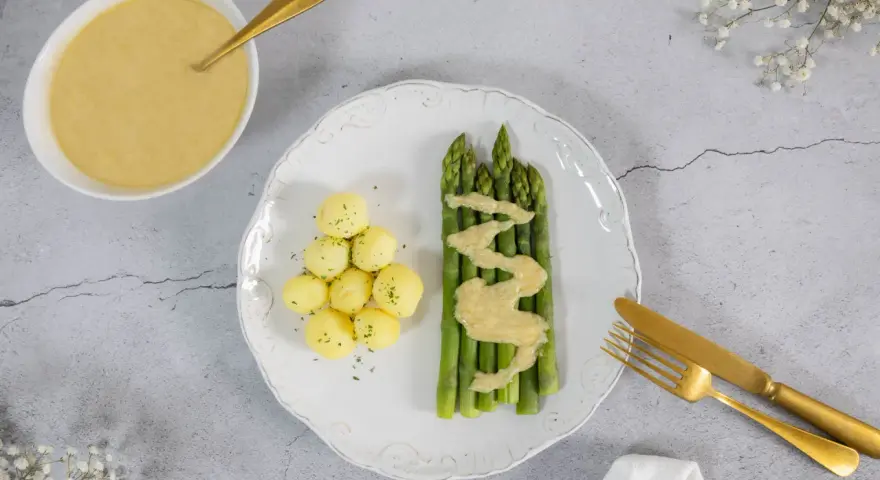 Spargel mit kleinen Kartoffeln und einer cremigen, veganen Sauce Hollandaise. Ein goldener Löffel liegt in der Sauce.