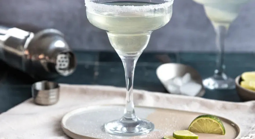 Ein perfekt zubereitetes Margarita-Rezept: Ein Glas mit Salzrand, daneben Limettenspalten, Eiswürfel und ein Shaker.