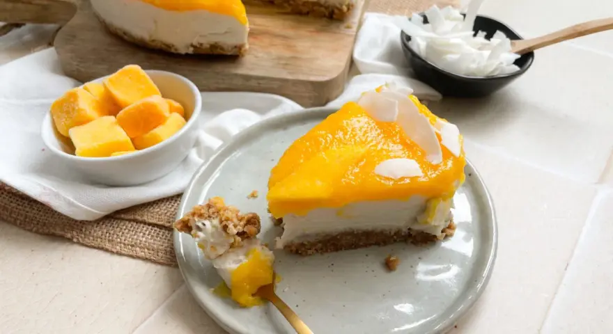 Ein cremiges Mango Cheesecake ohne Backen, garniert mit Kokosflocken. Ein Stück wird gerade mit einem Löffel probiert.