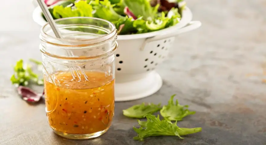 Ein Glas mit selbstgemachtem Salatdressing, das aussieht wie im Restaurant. Daneben ein Sieb mit frischem Salat.