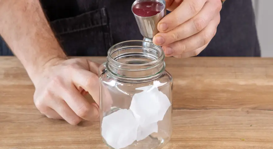 Eiswürfel in einem Glas, dazu wird rote Flüssigkeit aus einem Messbecher gegossen. So einfach lässt sich Rhabarberschorle selber machen!