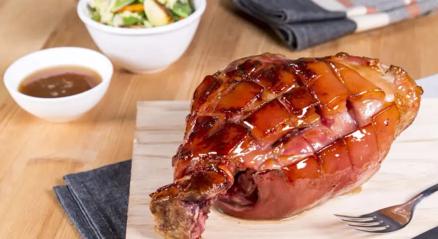 Glänzende, karamellisierte Schweinshaxe sous vide auf Holzbrett, daneben Schüssel mit Sauce und Salat.