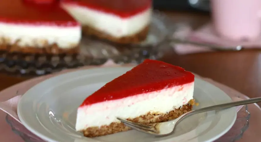 Ein Stück New York Cheesecake ohne Backen mit rotem Fruchttopping auf einem Teller, daneben ein ganzer Kuchen.