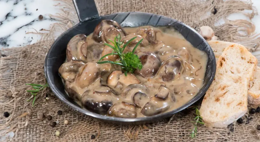 Herzhafte Champignon Rahmsauce in einer Pfanne, garniert mit Rosmarin, daneben knuspriges Brot und Pfefferkörner.