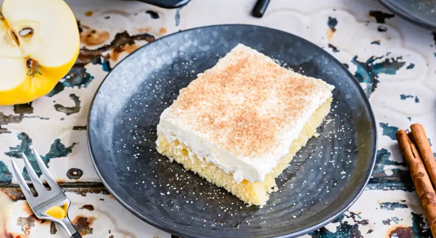 Ein Stück von Omas Apfelmuskuchen vom Blech, cremig belegt und mit Zimt bestreut, daneben ein halber Apfel und Zimtstangen.