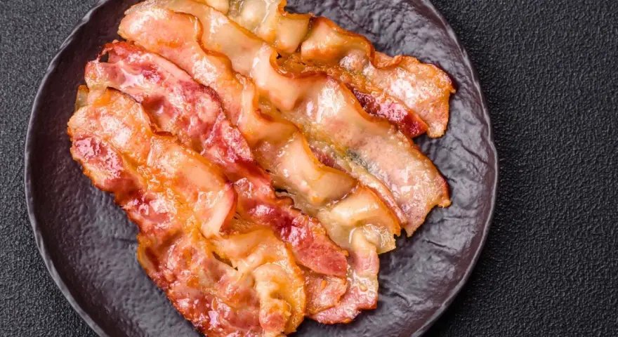 Knusprig gebratener Bacon im Ofen, auf einem dunklen Teller angerichtet.