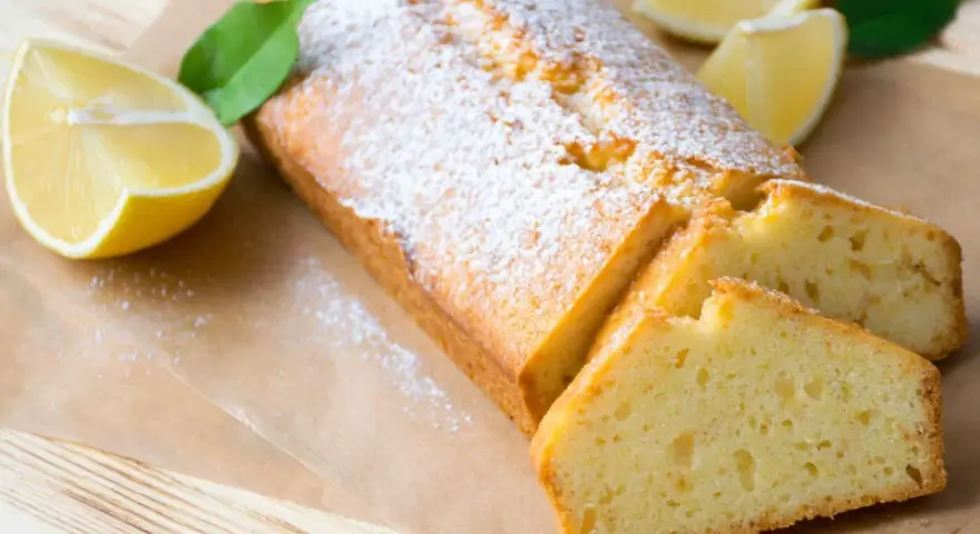 Saftiger veganer Zitronenkuchen, bestäubt mit Puderzucker, mit frischen Zitronenspalten und Blättern garniert.