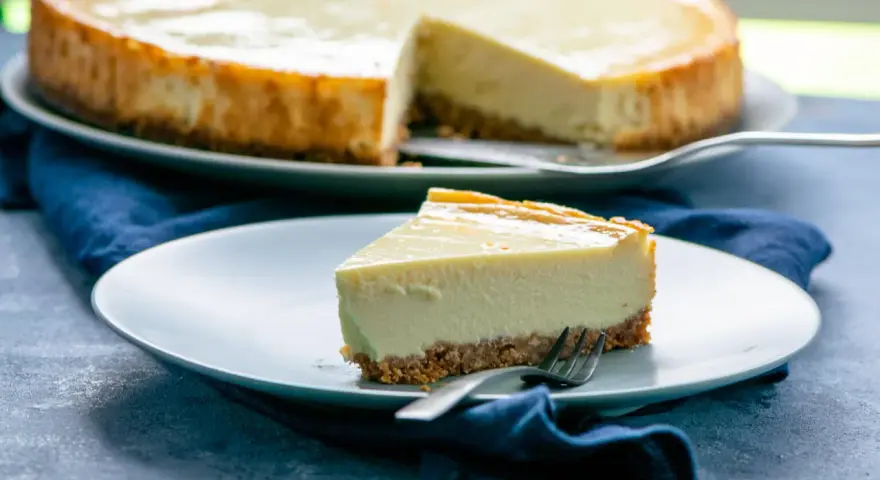Ein Stück cremiger american cheesecake mit knusprigem Boden auf einem Teller, daneben ein ganzer Kuchen.