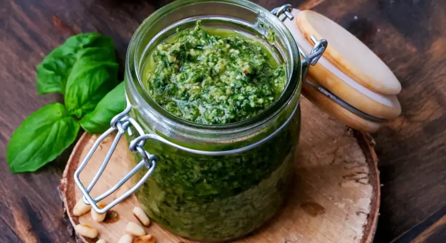 Frisches Basilikum Pesto in einem Glas, umgeben von Pinienkernen und Basilikumblättern.
