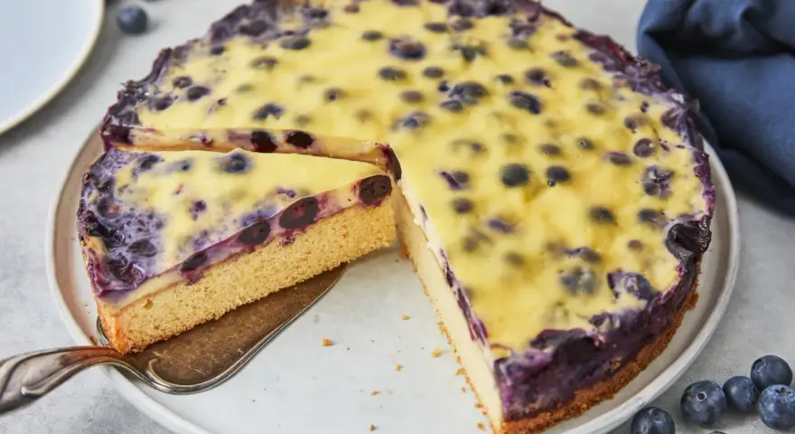 Ein saftiger Heidelbeerkuchen mit einer cremigen Vanillepudding-Schicht und frischen Heidelbeeren. Ein Stück wird gerade herausgeschnitten.
