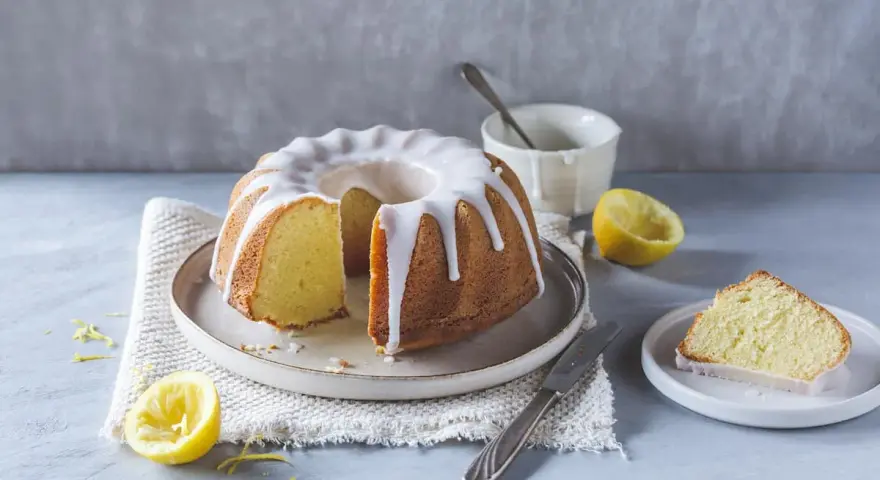 Ein saftiger Zitronenkuchen mit Zuckerguss, perfekt für ein Lemon Drizzle Cake Rezept. Daneben liegen frische Zitronen und ein Stück Kuchen.