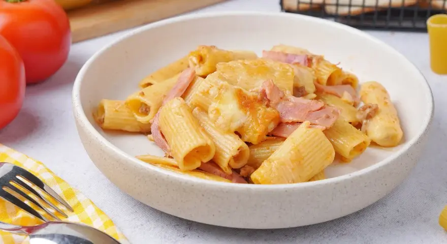 Herzhaftes Rigatoni al Forno mit Schinken und geschmolzenem Käse in einer Schüssel, daneben Tomaten und Brot.