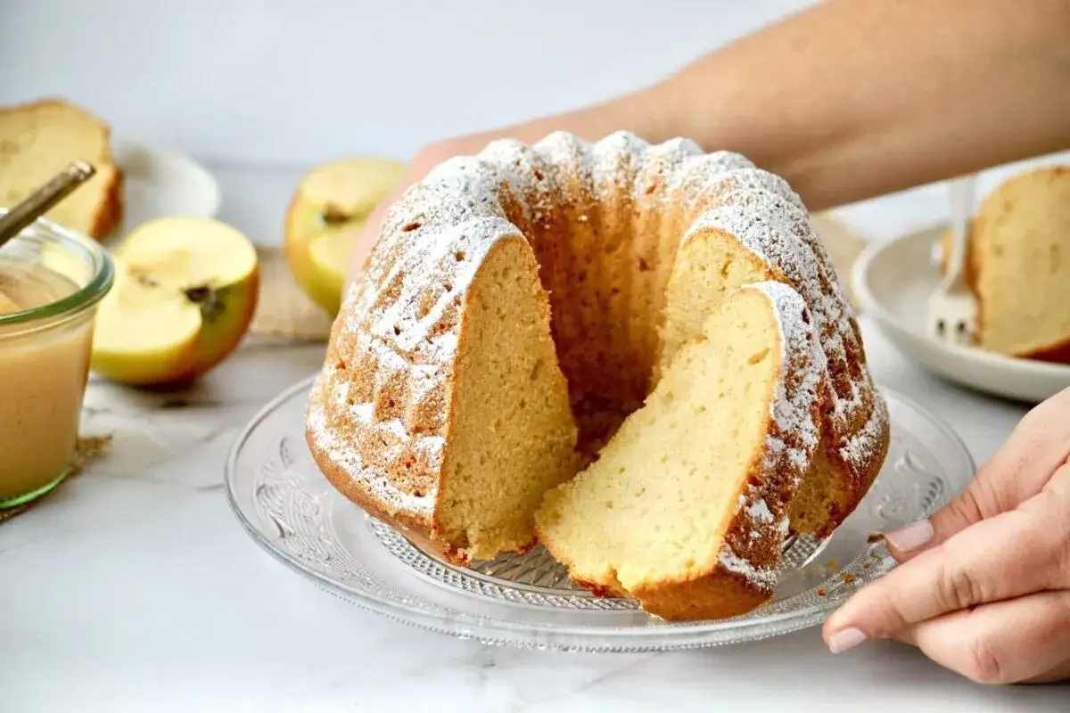 Ein schneller saftiger Kuchen mit Apfelmus wird auf einem Glasst&auml;nder pr&auml;sentiert, best&auml;ubt mit Puderzucker. Daneben liegen &Auml;pfel und ein Glas Apfelmus.