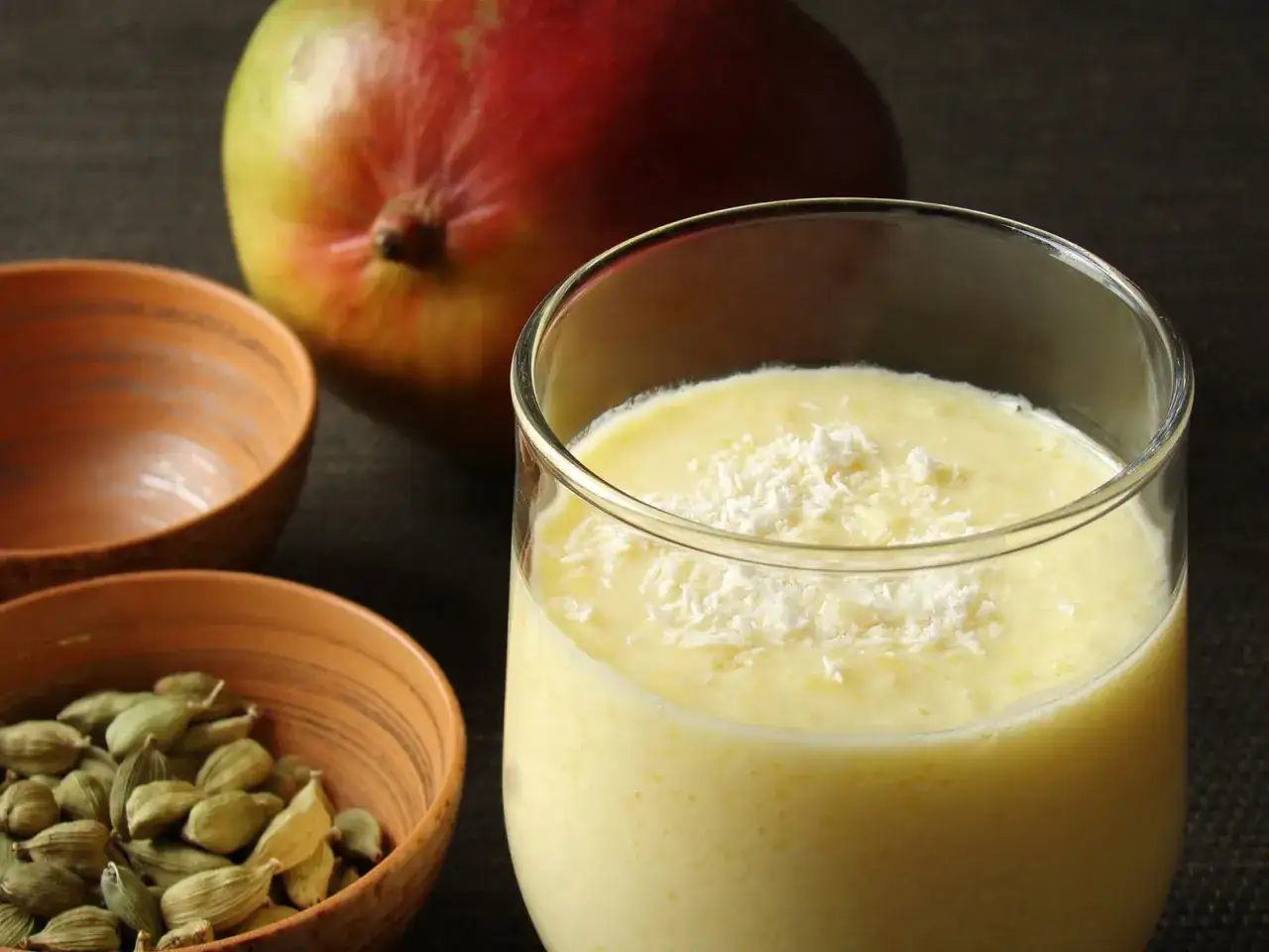 Cremiger Mango Lassi, garniert mit Kokosraspeln, neben einer reifen Mango und Kardamomkapseln in kleinen Schalen.