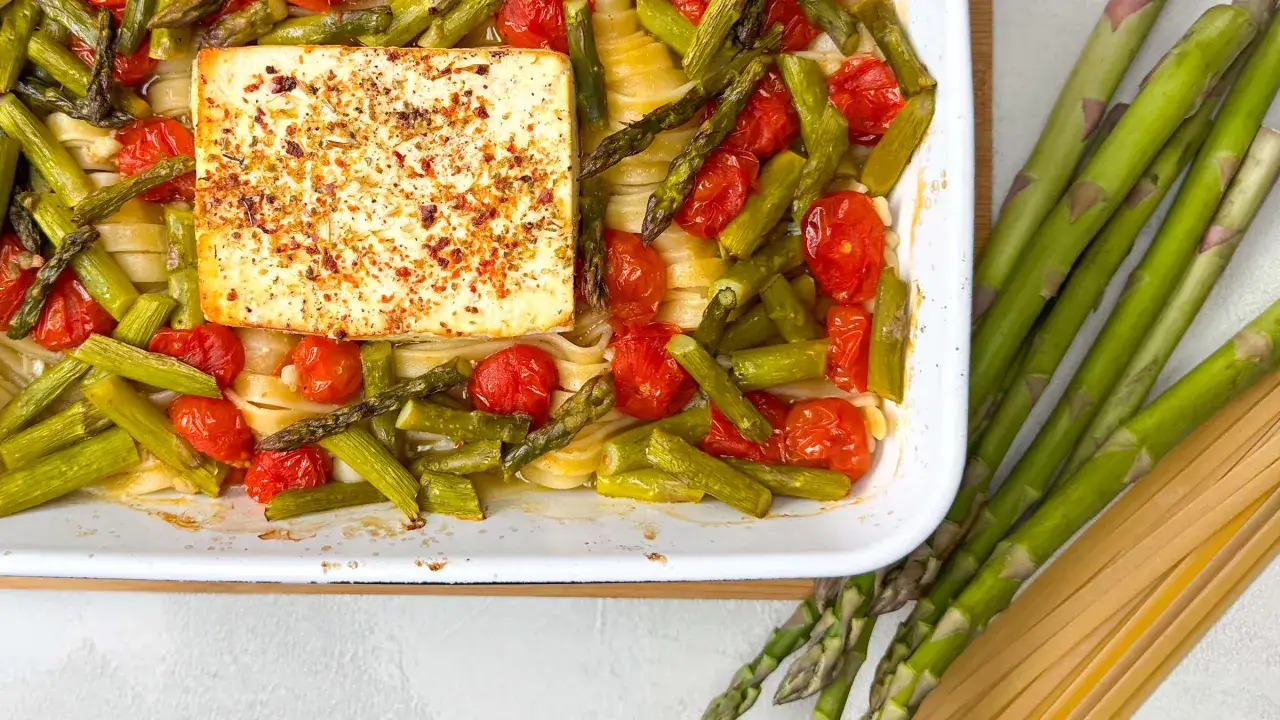 Eine Auflaufform mit Spargel, Kirschtomaten und Pasta, gekr&ouml;nt von einem Block Feta. Daneben liegen frischer Spargel und ungekochte Pasta.