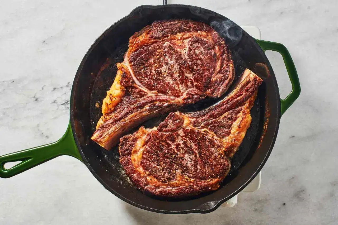 Zwei saftige Rib-Eye-Steaks braten in einer Gusseisenpfanne. Rauch steigt auf, während sie perfekt gegart werden.