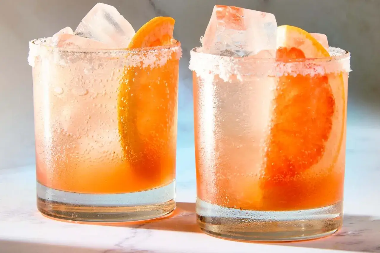 Zwei erfrischende Aperol Paloma Cocktails mit Grapefruitspalten und Eis, die R&auml;nder der Gl&auml;ser sind mit Salz verziert.