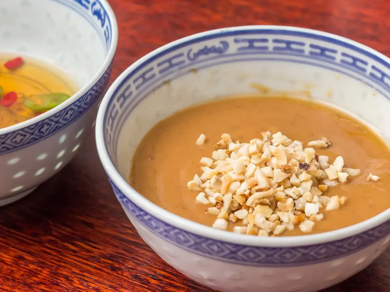 Cremige Erdnuss Sauce selber machen, garniert mit gehackten Erdn&uuml;ssen in einer Sch&uuml;ssel mit blau-wei&szlig;em Muster.